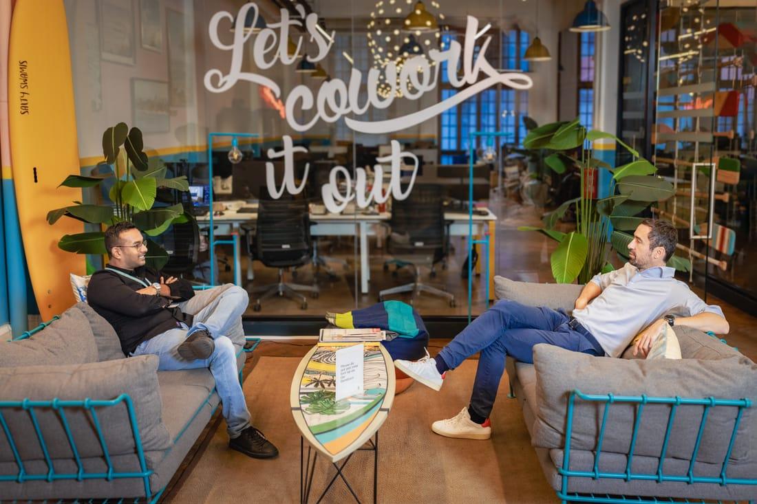 Two colleagues on sofas beneath a neon sign reading Let's cowork it out
